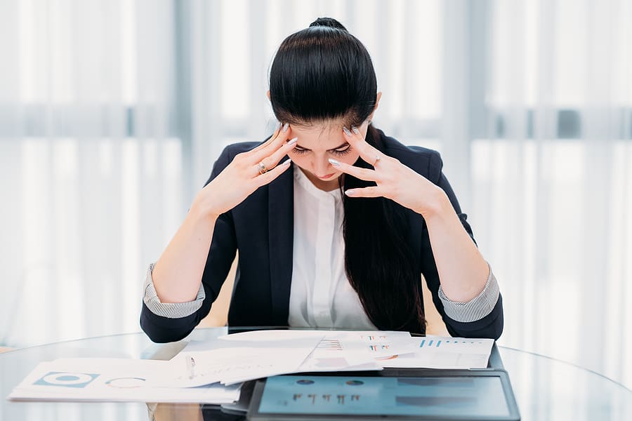 Woman sitting at a table, holding her head in her hands and looking overwhelmed at the papers in front of her.